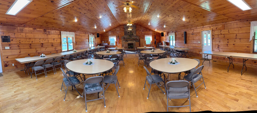 Spacious event hall at Keith Memorial Park featuring round tables, wooden walls, and a central stone fireplace.